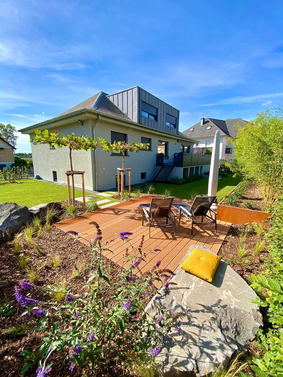Modernes Haus mit Terrasse, stilvollem Outdoormöbel, Loungesesseln und einem Garten mit einem Kissen auf einem Felsen unter einem klaren blauen Himmel. Ein perfektes Beispiel für zeitgenössisches Gardendesign in Deutschland.