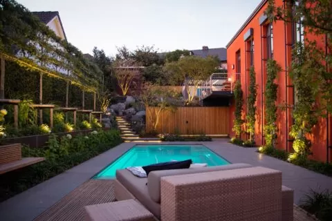Moderner Garten mit beleuchtetem Pool, stilvollen Outdoormöbeln, Liegestühlen, üppigem Grün und einem leuchtend orangefarbenen Gebäude in der Abenddämmerung.