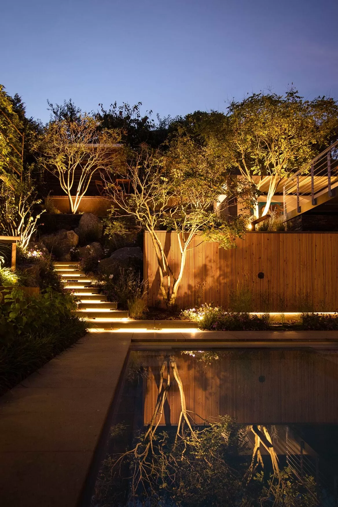 Der Garten mit beleuchteten Stufen, Bäumen, stilvollen Outdoormöbeln und Holzzaun spiegelt sich in einem ruhigen Pool in der Abenddämmerung.