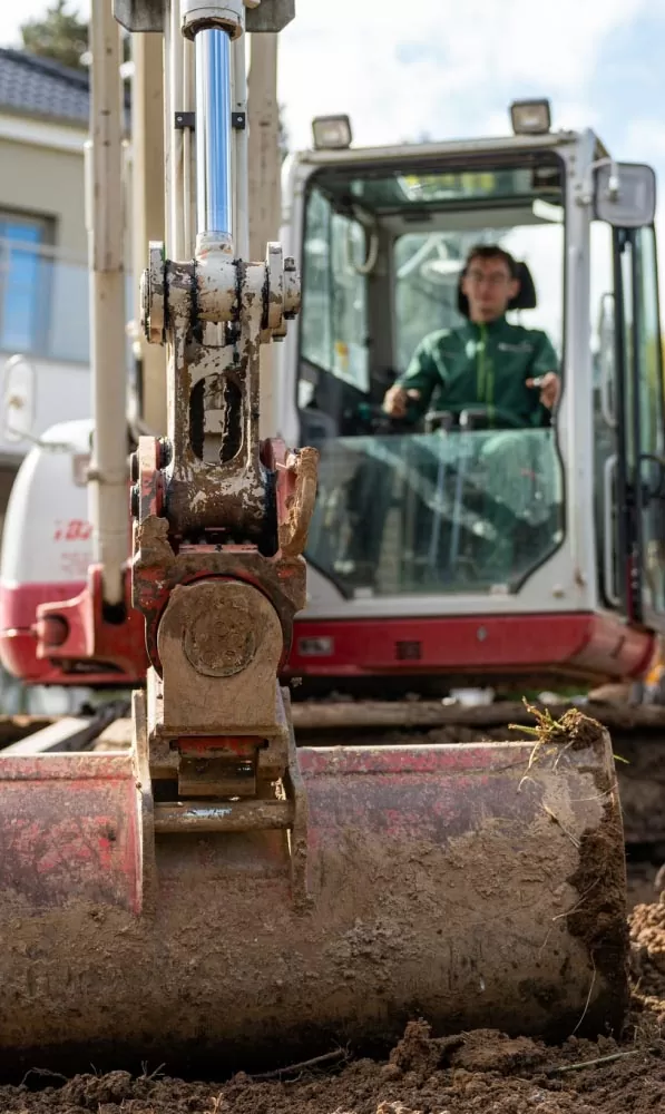 Eine Person bedient einen kleinen Bagger für den Gartenbau, wobei der Schwerpunkt auf der Schlammschaufel und dem Hydraulikarm liegt.
