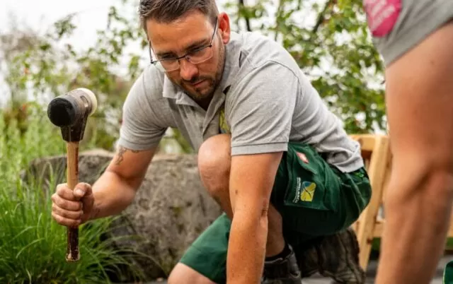 Ein Mann mit Brille kniet in Luxemburg im Freien, hält einen Hammer in der Hand und arbeitet an einem Outdoormöbel-Projekt.