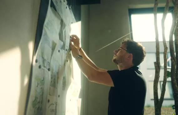 Ein Mann mit Brille und schwarzem Hemd heftet in einem sonnendurchfluteten Raum, in dem große Fenster und ein Ast im Hintergrund zu sehen sind, Papiere an eine Tafel. Er plant Garten- und Landschaftsbauprojekte für Burelbach in Luxemburg und Deutschland.