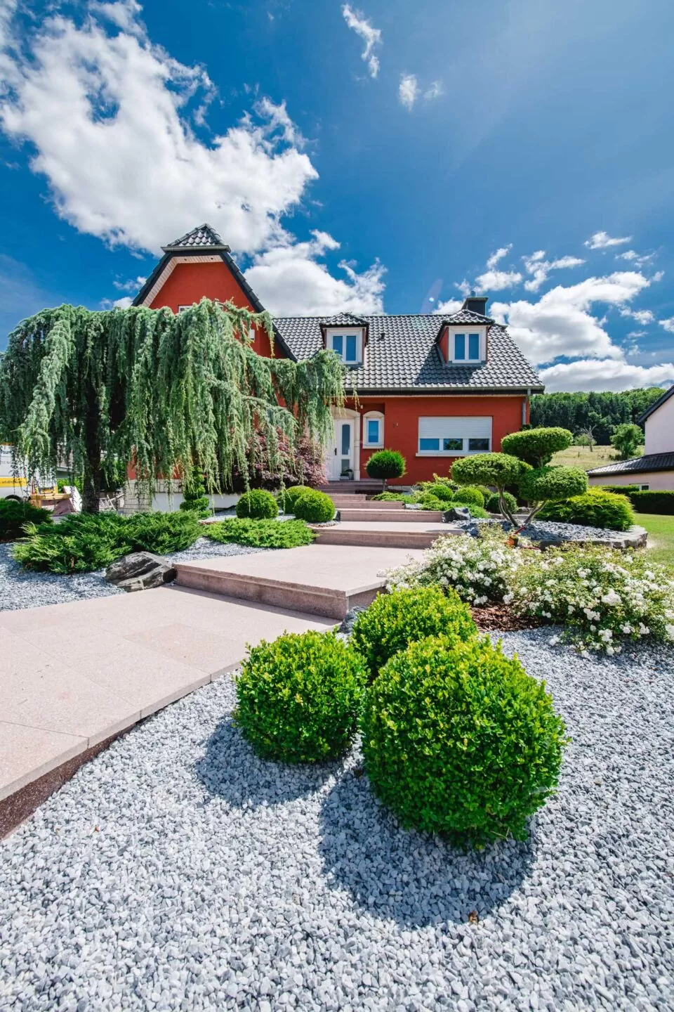 Ein rotes Haus mit gepflegten Sträuchern von Burelbach, ein steinerner Gehweg und ein strahlend blauer Himmel mit vereinzelten Wolken über dem Kopf - das ist eleganter Gartenbau in Luxemburg.