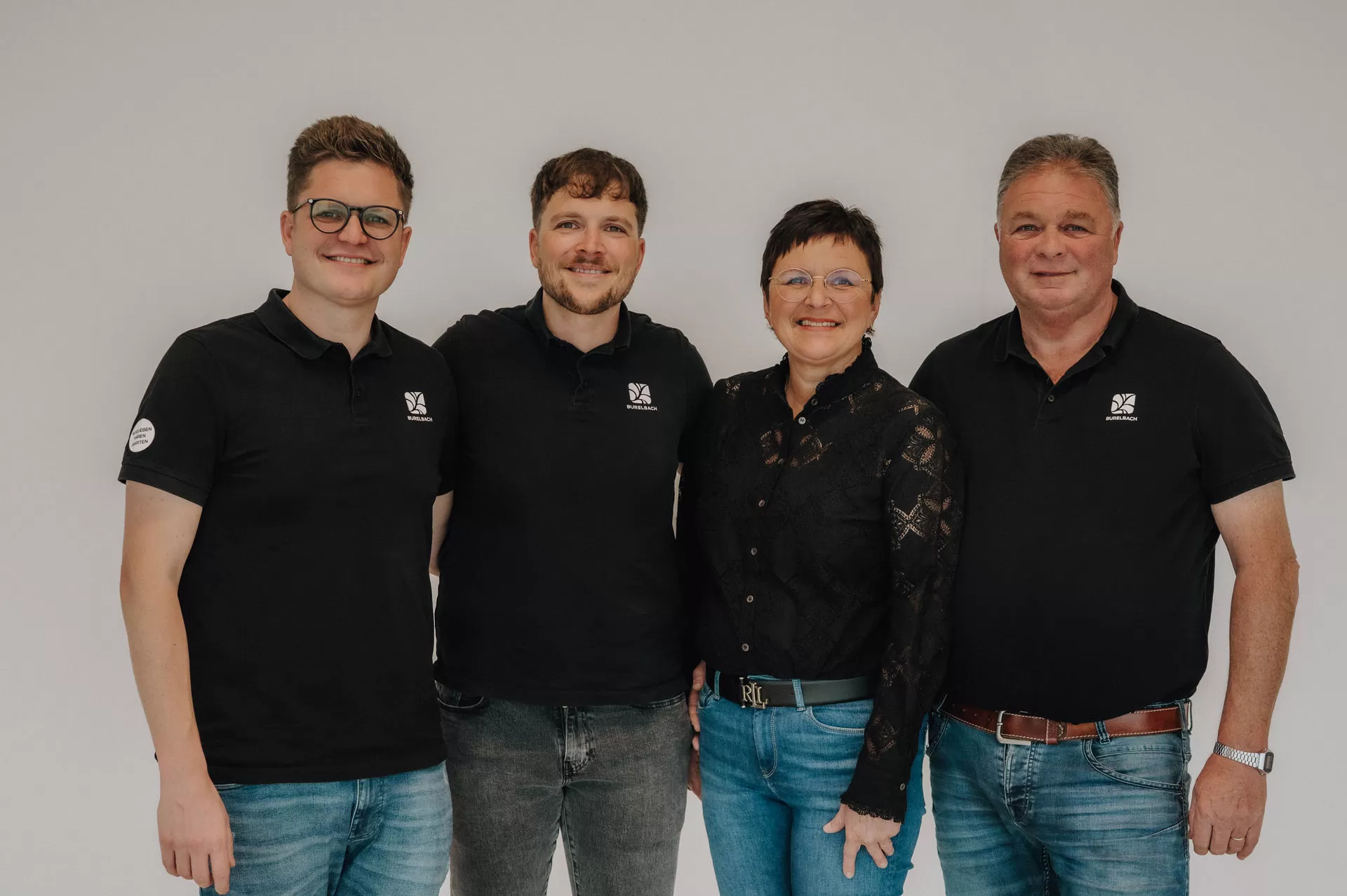 Familie Burelbach in schwarzen Oberteilen stehen lächelnd nebeneinander vor einem schlichten hellen Hintergrund und zeigen ihre Leidenschaft für Gardendesign und Outdoormöbel.
