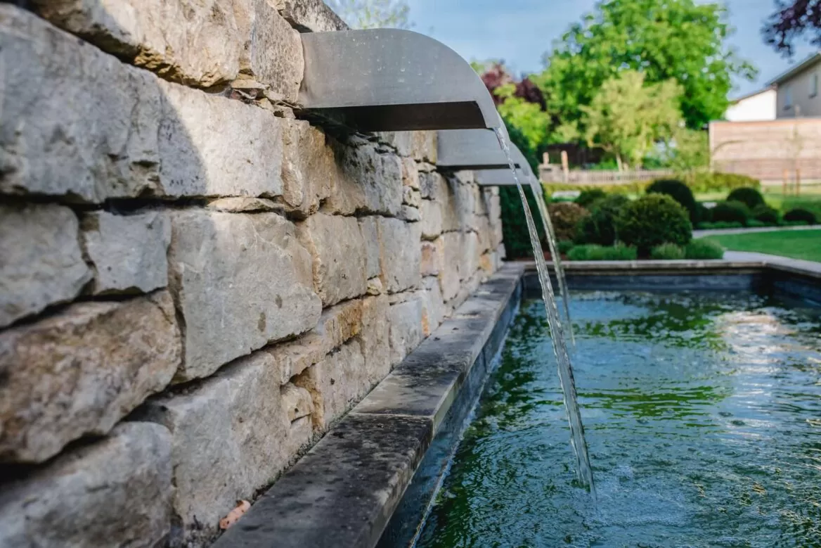 Wasser fließt aus einem Metallauslauf an einer Steinmauer in einen rechteckigen Gartenteich, umgeben von Grünzeug und stilvollen Outdoormöbeln von Burelbach, Deutschland.