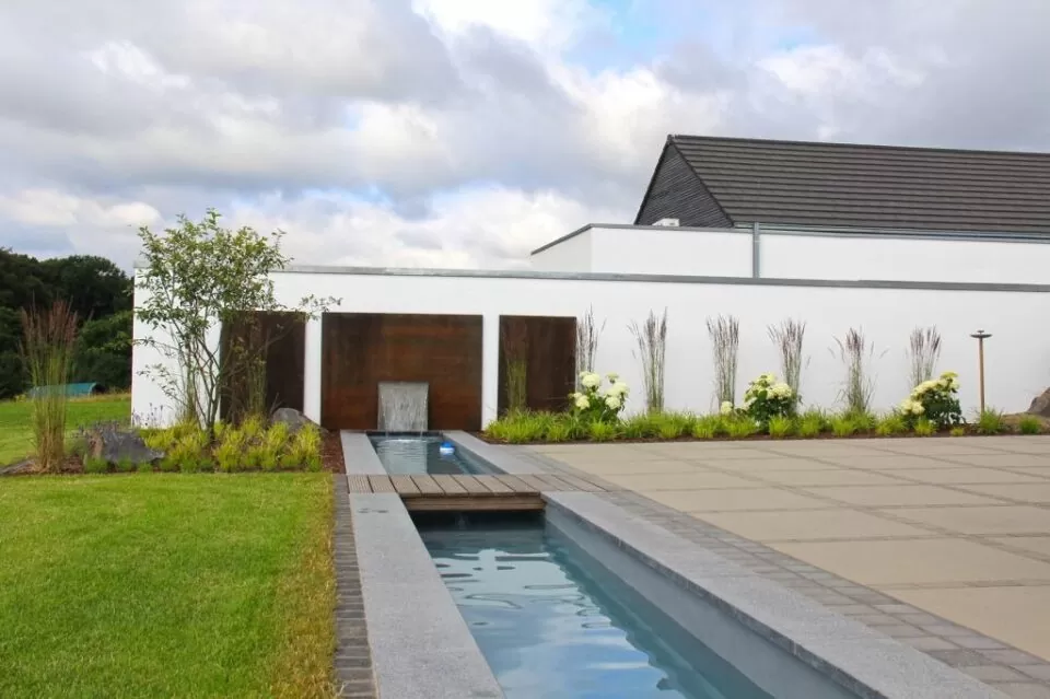 Moderner Garten mit Outdoormöbeln, einem schmalen Wasserspiel, einer weißen Mauer, stilvollen Pflanzen und einer sauber gepflasterten Terrasse unter einem wolkenverhangenen Himmel - perfekt für zeitgenössisches Gardendesign in Luxemburg.