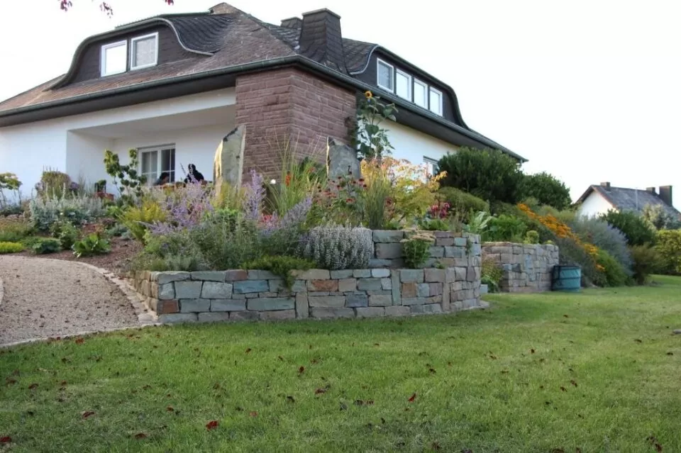 Ein Haus in Burelbach mit einer steinernen Stützmauer, bunten Gartenbeeten und einem grünen Rasen davor.
