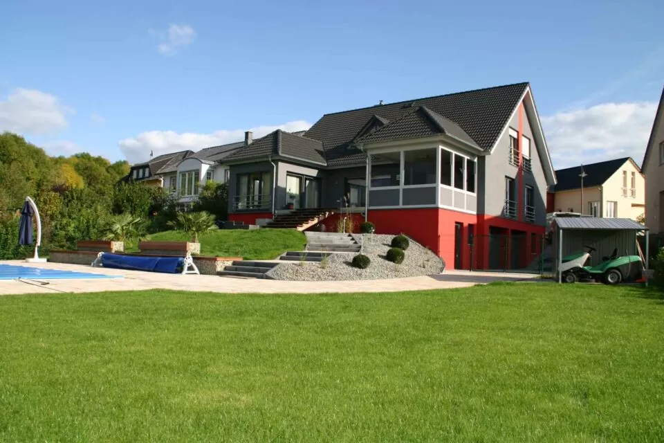 Modernes Haus mit großen Fenstern, roten Akzenten und grünem Rasen in Deutschland; Outdoormöbel auf der Terrasse, mit Pool und Garten im Vordergrund.