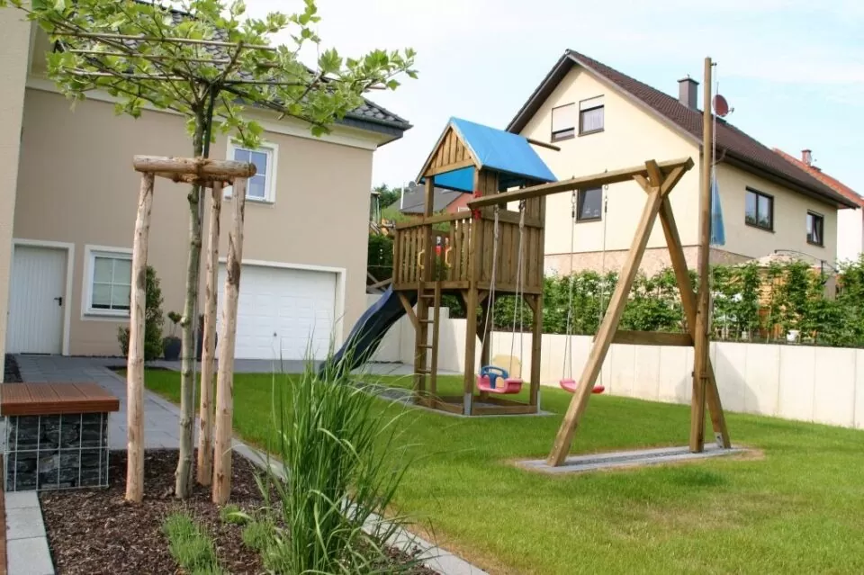 Holzspielplatz mit Schaukeln und Rutsche in einem grasbewachsenen Gartenbau-Hinterhof in der Nähe eines modernen Hauses und eines kleinen Baumes in Deutschland.