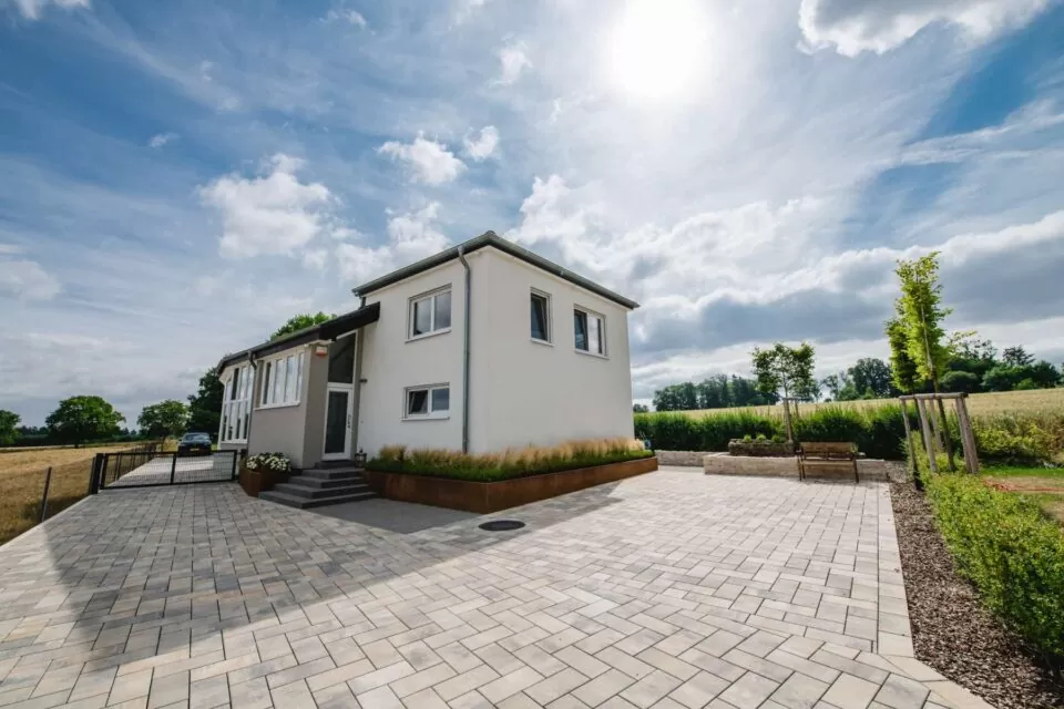 Modernes weißes Haus mit großer gepflasterter Auffahrt, ergänzt durch stilvolle Outdoormöbel und umgeben von grünen Feldern unter einem hellen, sonnigen Himmel.
