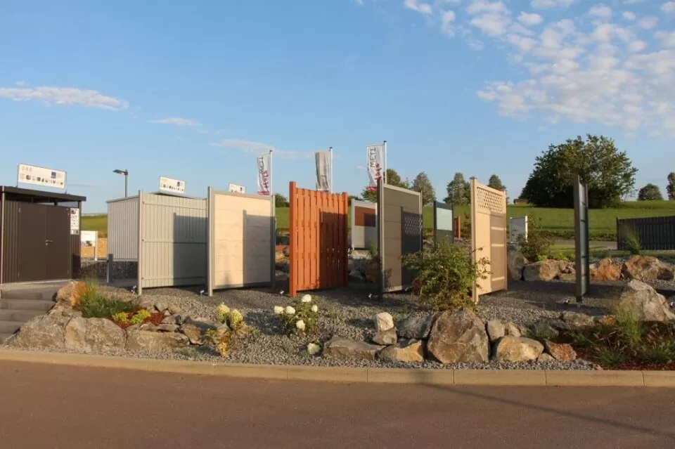 Ausstellung verschiedener Zaun- und Torpaneele im Freien auf Kies, umgeben von Landschaftsgestaltung und Felsen, unter blauem Himmel - ein Beweis für die Kompetenz von Burelbach im Gartendesign.