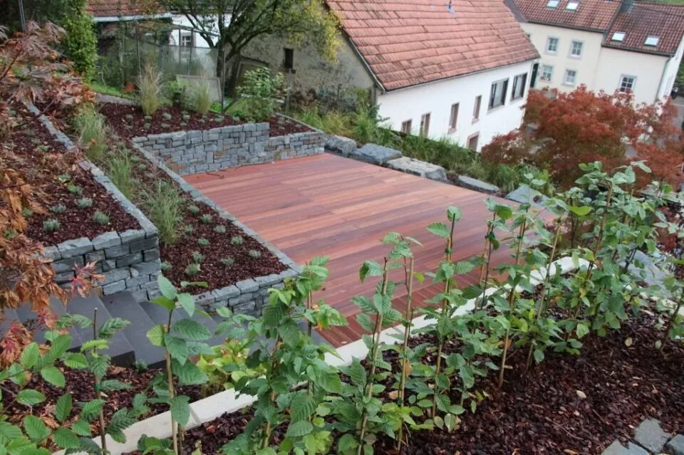 Mehrstöckiger Garten mit Holzdeck, Steinmauern und Grünpflanzen, mit stilvollen Outdoormöbeln, neben Häusern mit roten Dächern. Ein perfektes Beispiel für modernes Gardendesign in Deutschland.