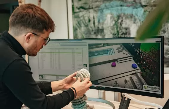Ein Mann mit Brille arbeitet an einem Schreibtisch und untersucht ein aufgerolltes Objekt, während er auf einen Computermonitor blickt, auf dem ein 3D-Modell und eine Software für Gartengestaltung zu sehen sind, die modernes Handwerk in Deutschland und Luxemburg zeigen.