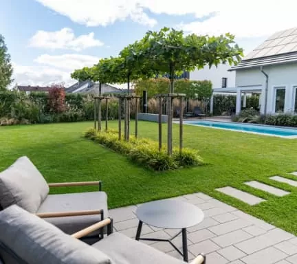 Moderner Garten mit stilvollen Gartenmöbeln auf einem Terrassensitzplatz, einem kleinen runden Tisch, ordentlich getrimmtem Rasen, einer Reihe junger Bäume, Trittsteinen und einem Swimmingpool - perfektes Gardendesign für Deutschland und Luxemburg.