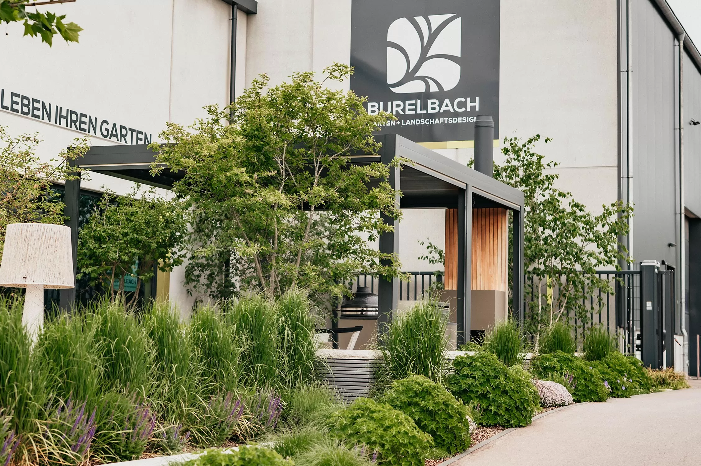 Moderne Gartengestaltung mit üppigen Grünpflanzen, hohen Gräsern und einem schattigen Sitzbereich vor einem Gebäude mit einem "Burelbach"-Schild. Die Umgebung wirkt sauber und gepflegt.